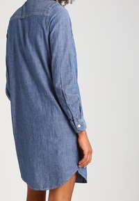 Abito camicia in denim di blu chiaro, con colletto, maniche lunghe, polsini con bottoni e orlo arrotondato con spacchi laterali per facilitare i movimenti.