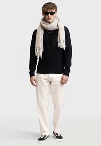 Mørkeblå strikket sweater med lomme, cremefarvet tørklæde, cremefarvede bukser og sorte sneakers. Modellen bærer solbriller og står foran en ensfarvet baggrund.