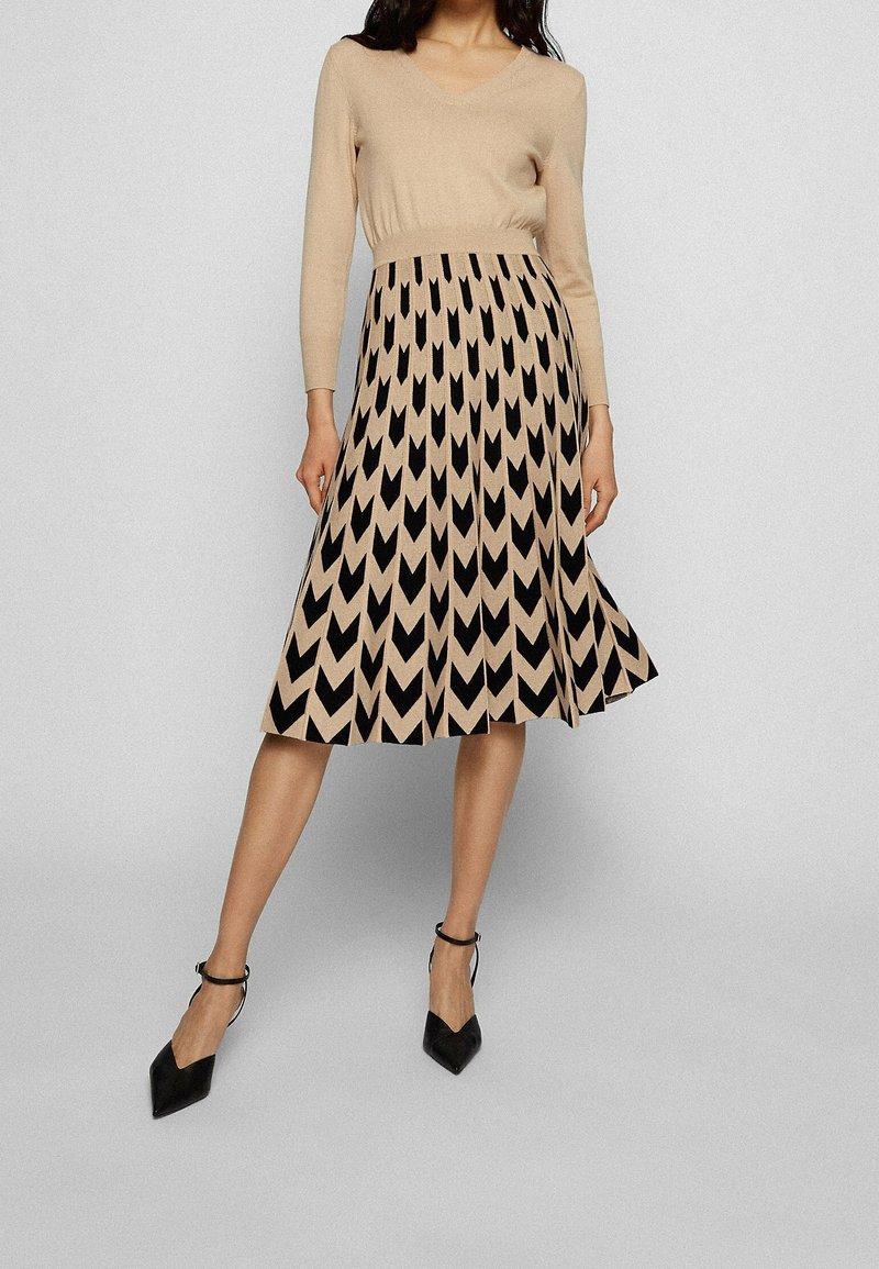Femme portant un pull beige à col en V associé à une jupe midi plissée beige et noire à motif, et des talons noirs à bride à la cheville.