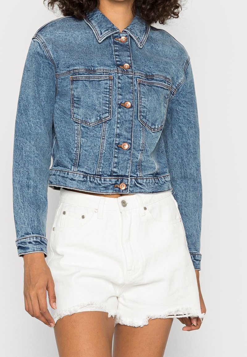 Blue denim cropped jacket with copper buttons, two front pockets, and contrasting stitching, paired with white distressed shorts.