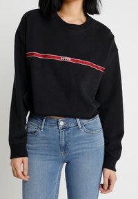 Sweat-shirt noir avec une bande horizontale rouge et blanche portant le texte "Levi's", associé à un jean bleu clair, coupe décontractée.
