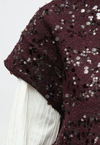 Burgundska teksturirana tkanina s crnim sjajnim detaljima postavljena preko bijele plisirane tkanine na rukavu odjeće.