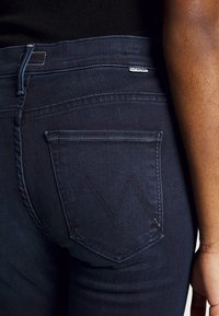 Mörkblå denimjeans med en närbild av bakfickan, som har ett sydda chevronmönster och en slät yta.