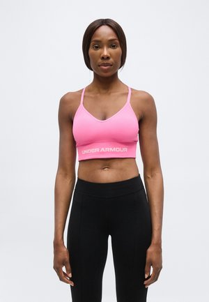 Kobieta stoi zwrócona przodem, ubrana w różowy sportowy biustonosz Under Armour i czarne legginsy z wysokim stanem na jednolitym tle.