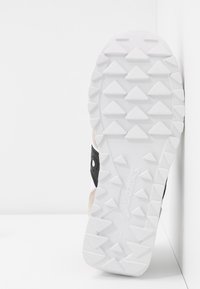 Semelle d'une chaussure de sport blanche avec un motif de bande de roulement triangulaire et un logo noir sur le côté, positionnée verticalement contre un mur blanc.