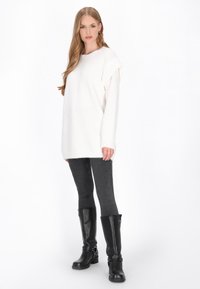 Túlságosan nagy, fehér kötött pulóver, puha textúrával, sötétszürke leggings-szel és fekete térdimmärci csizmával, amelyeken csatok találhatók.