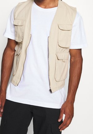 Homme portant un gilet utilitaire beige avec plusieurs poches par-dessus un t-shirt blanc et un pantalon noir, montré des épaules aux hanches.