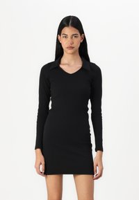 Μαύρο φόρεμα μακρυμάνικο με εφαρμοστή γραμμή, V-λαιμόκοψη και μυτερό γιακά. Φτιαγμένο από λείο ύφασμα, φτάνει μέχρι το μεσοφόρι.