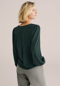 Blusa verde scuro con un tessuto morbido e textured, scollatura rotonda e maniche a palloncino. Include un dettaglio con una piega sul retro, abbinata a pantaloni grigi.
