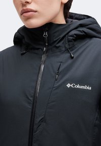 Άτομο που φοράει μαύρο μπουφάν Columbia με μπροστινό φερμουάρ και κουκούλα, δείχνοντας το λογότυπο στο στήθος.