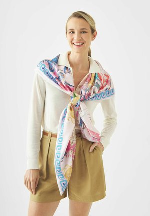 Glimlachende vrouw die een witte trui en lichtbruine korte broek draagt, met een kleurrijk sjaal met patroon om haar schouders geknoopt, hand in de zak.