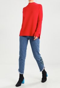Red ribbed sweater with a high neckline, paired with light blue jeans featuring a green patterned accent and frayed hem, complemented by black ankle boots with blue block heels.