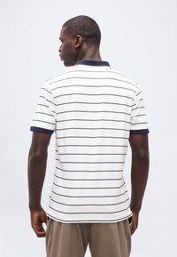 Άνδρας που φοράει λευκή κοντομάνικη πόλο μπλούζα με λεπτές μαύρες οριζόντιες ρίγες και ναυτικό γιακά, στραμμένος προς τα πίσω μπροστά σε απλό φόντο.