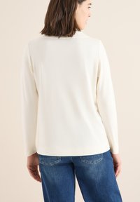 Long-sleeve, cream-colored top with a smooth texture, paired with blue jeans featuring subtle stitching and back pockets.