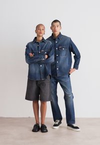 To modeller har på seg denimantrekk: den ene har på seg en mørk denimskjorte og shorts, mens den andre har på seg en lysere denimjakke og jeans. Begge har sorte sko.