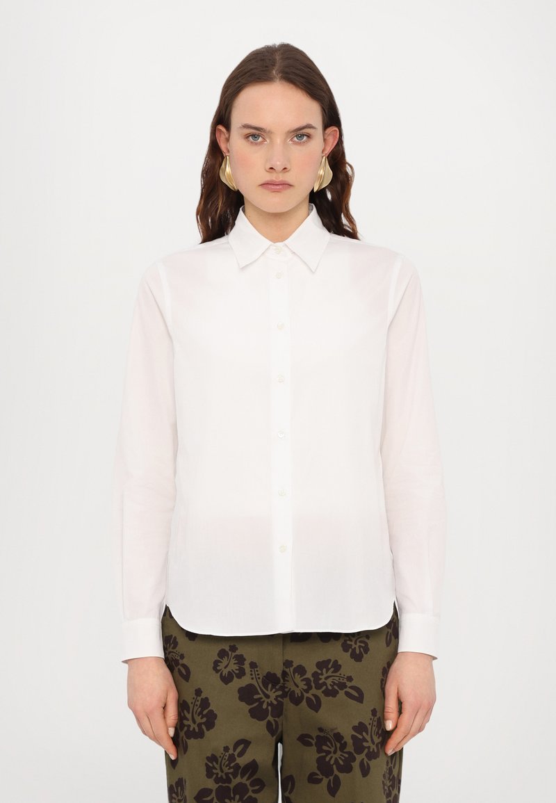 Chemise blanche à manches longues avec un col pointe et fermeture à boutons, fabriquée dans un tissu lisse, associée à un pantalon à motif floral sombre.