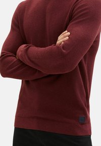Bordeauxfarbener Strickpullover mit strukturiertem Muster, Rundhalsausschnitt und gerippten Bündchen. Verfügt über ein kleines Logo-Patch nahe dem Saum.