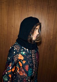 Kapucnis fekete ruhadarab leopárdmintás belsővel, amely egy színes virág- és állatmintás dzsekivel van párosítva. Texturált szövet részletek láthatók.