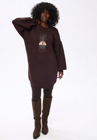 Bruine oversized sweaterjurk met een boomgrafiek en wortels, met lange mouwen en een geribde zoom. Gecombineerd met bruine legging en kniehoge laarzen.