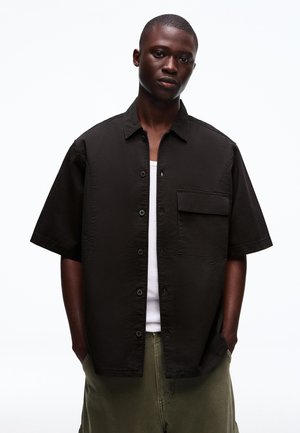 Junger Mann mit kurzen Haaren steht mit den Händen in den Taschen, trägt ein schwarzes kurzärmeliges Hemd über einem weißen Tanktop und olivfarbene Hosen.