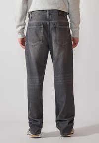 Dark grey denim jeans with a relaxed fit, featuring back pockets, visible stitching, and a branded waistband. Tapered leg design.