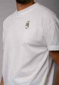 Weißes Baumwoll-T-Shirt mit einem kleinen grünen Schlangen-Stickerei auf der linken Brust. Kurze Ärmel mit umgeschlagenen Säumen, lässige Passform.