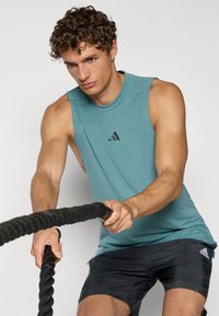 Søblå ærmeløs træningstop med rund halsudskæring og sort Adidas-logo, iført med sorte shorts, mens en person griber om kampe reb.