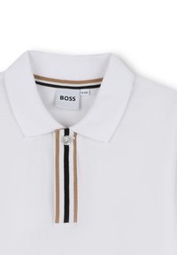 Vit polo skjorta med texturerat tyg, med en treknappslå och en krage med bruna, svarta och vita ränder. Pr präglad logotyp.