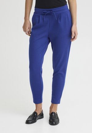 Kobieta ubrana w niebieskie spodnie na sznurek i czarne mokasyny, stojąca na tle jasnego, jednolitego tła, ręce swobodnie opuszczone wzdłuż ciała.