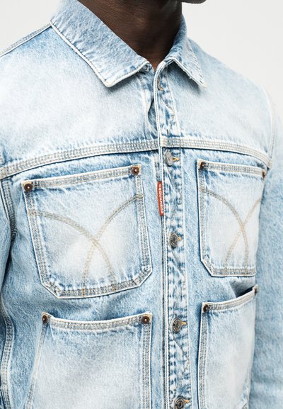 Ανοιχτό μπλε τζιν μπουφάν με τ έσσερις μπροστινές τσέπες, μεταλλικά κουμπιά και εμφανή ραφές, που φοριέται από άτομο με σκουρόχρωμο δέρμα.