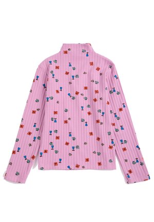 Fille portant un col roulé fleur rose, un pantalon corail à jambes larges, des chaussures noires et un bandeau tressé rouge, blanc et bleu, debout sur un fond uni.