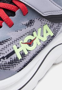 Κοντινό πλάνο ενός γκρι και μαύρου αθλητικού παπουτσιού με κόκκινες κορδόνια, λευκή σόλα και λογότυπο HOKA σε ανοιχτό πράσινο με κόκκινη λεπτομέρεια στο πλάι.