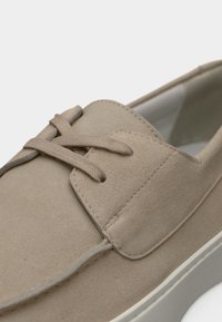 Chaussure en daim beige à texture lisse, avec un design à lacets, un bout arrondi et une semelle en caoutchouc blanche. Présente des coutures discrètes et un col rembourré.