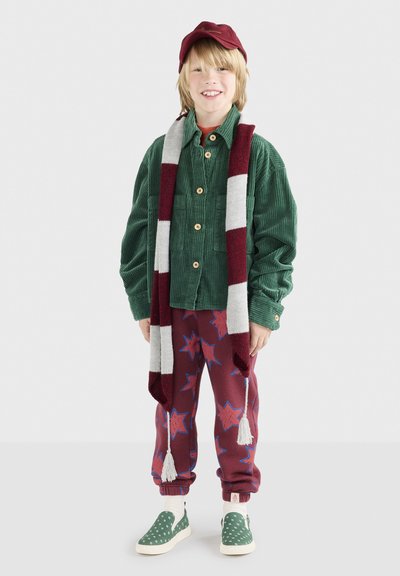 Giacca verde in corduroy con bottoni di legno, sciarpa a righe in maglia, pantaloni della tuta bordeaux a fantasia con stelle e scarpe slip-on verdi a pois.