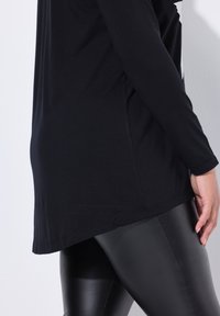 Zwarte langmouwtop met een ronde zoom en zijsplit, gecombineerd met glanzende zwarte leggings. Stof lijkt zacht en rekbaar te zijn.