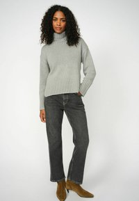 Grijze gebreide trui met een hoge col en geribbelde zoom, gecombineerd met donkere jeans en bruine enkellaarsjes. Overal een gladde textuur.