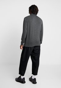 Pull en gris à manches longues avec un col haut, porté avec un pantalon noir ample et des chaussures de sport noires, affichant une coupe décontractée et relaxée.