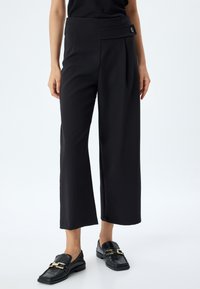 Zwarte wijde broek met een hoge taille en zijplooien, voorzien van een knoopdetail bij de taille. Gemaakt in combinatie met gestructureerde zwarte loafers.