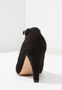 Svart mocka högklackad sko med böjd klack och ankelsnodd. Har en slät yta och en beige innersula, som visar en klassisk design.