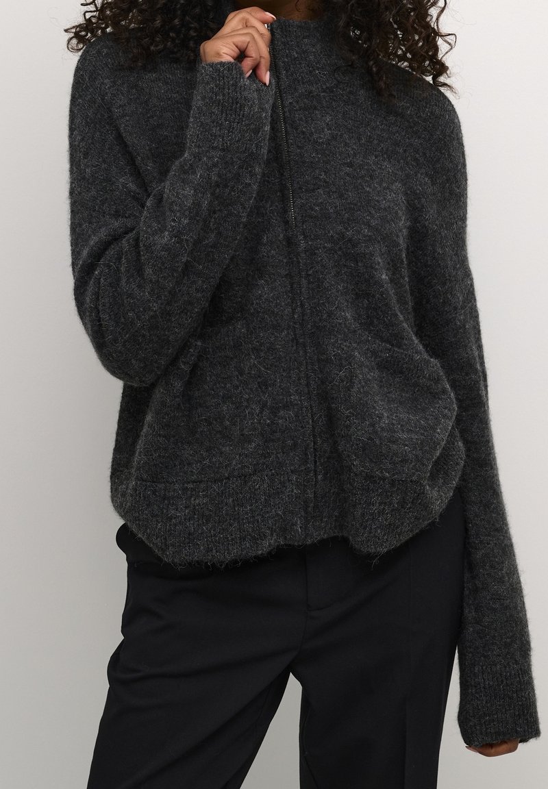 Femme portant un pull à fermeture éclair gris foncé en tissu pelucheux à manches longues et un pantalon noir, main près du col zippé sur un fond uni.