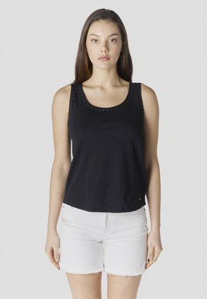 Femeie cu păr lung și castaniu, purtând un top negru fără mâneci și pantaloni scurți albi din denim, stând în picioare pe un fundal simplu, deschis la culoare.