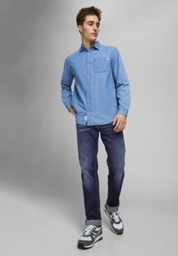 Giovane uomo che indossa una camicia di denim blu, jeans blu scuro con risvolti e sneakers verdi e bianche, in piedi contro uno sfondo grigio uniforme.