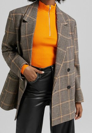 Personne portant un blazer marron à carreaux sur un pull zippé côtelé orange vif et un pantalon en cuir noir avec une main dans la poche.