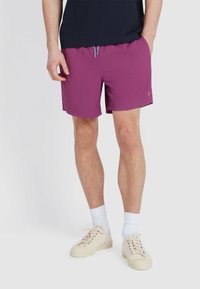 Paarse zwembroek met elastische tailleband, blauwe trekkoord en klein logo detail. Gedragen met witte crew sokken en beige sneakers.