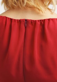 Il tessuto del vestito rosso presenta un collo arricciato e una cerniera nascosta sul retro. Il materiale sembra leggero e dalla texture interessante.