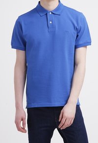 Camisa polo de algodón azul con cuello, tapeta de dos botones y mangas cortas; presenta un pequeño logo en el lado izquierdo.