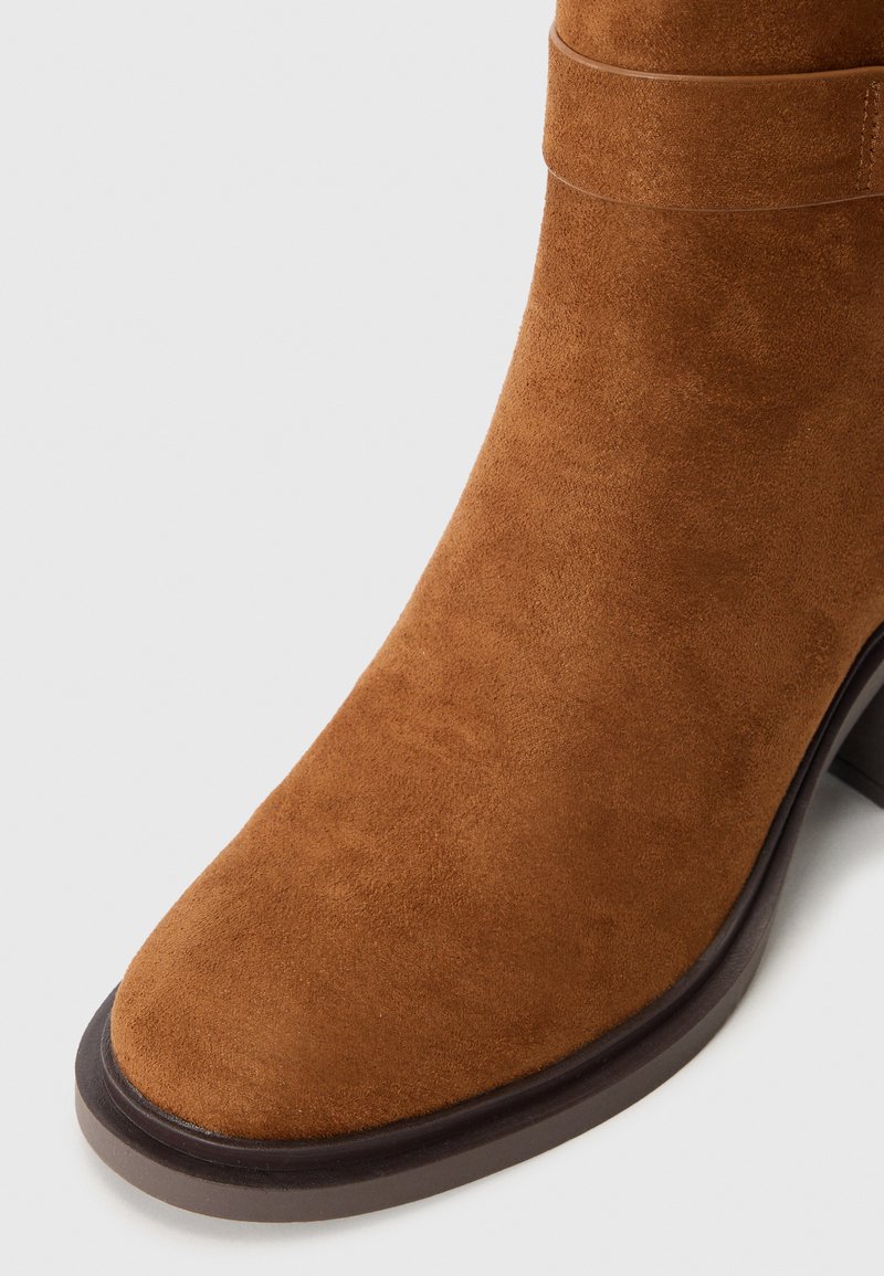 Botín de ante marrón hasta el tobillo con punta redonda y tacón robusto. Presenta un detalle de correa simple alrededor del tobillo y una suela lisa.