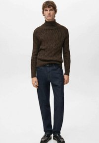 Brązowy golf z dzianiny na grubym splocie w połączeniu z ciemnoniebieskimi dżinsami. Model nosi czarne buty, stoi na neutralnym tle.