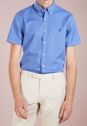 Mann trägt ein hellblaues, kurzärmeliges Hemd, das in beige Hosen gesteckt ist, vor einem einfarbigen Hintergrund.