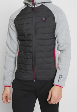 Mężczyzna w czarnej pikowanej kurtce z szarymi raglanowymi rękawami, czerwonymi akcentami zamka błyskawicznego oraz napisem "J&J Core 1990" na lewym przedramieniu.